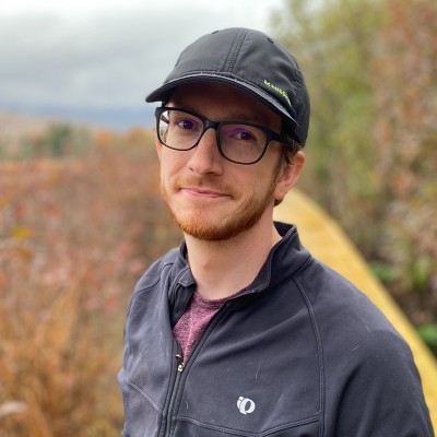 Headshot of Justin wearing a black hat and glasses in a wooded scene.